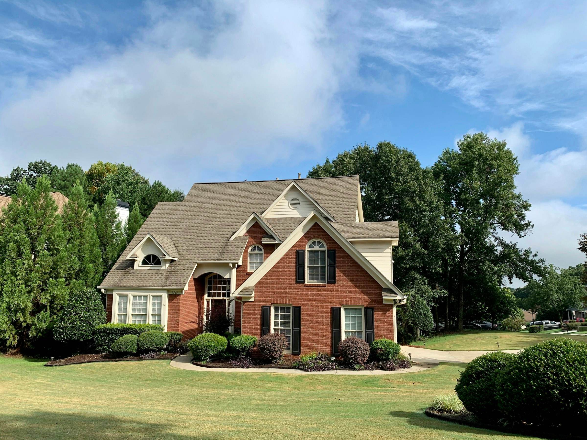 A two-story brick home with a steep shingle roof and trimmed hedges