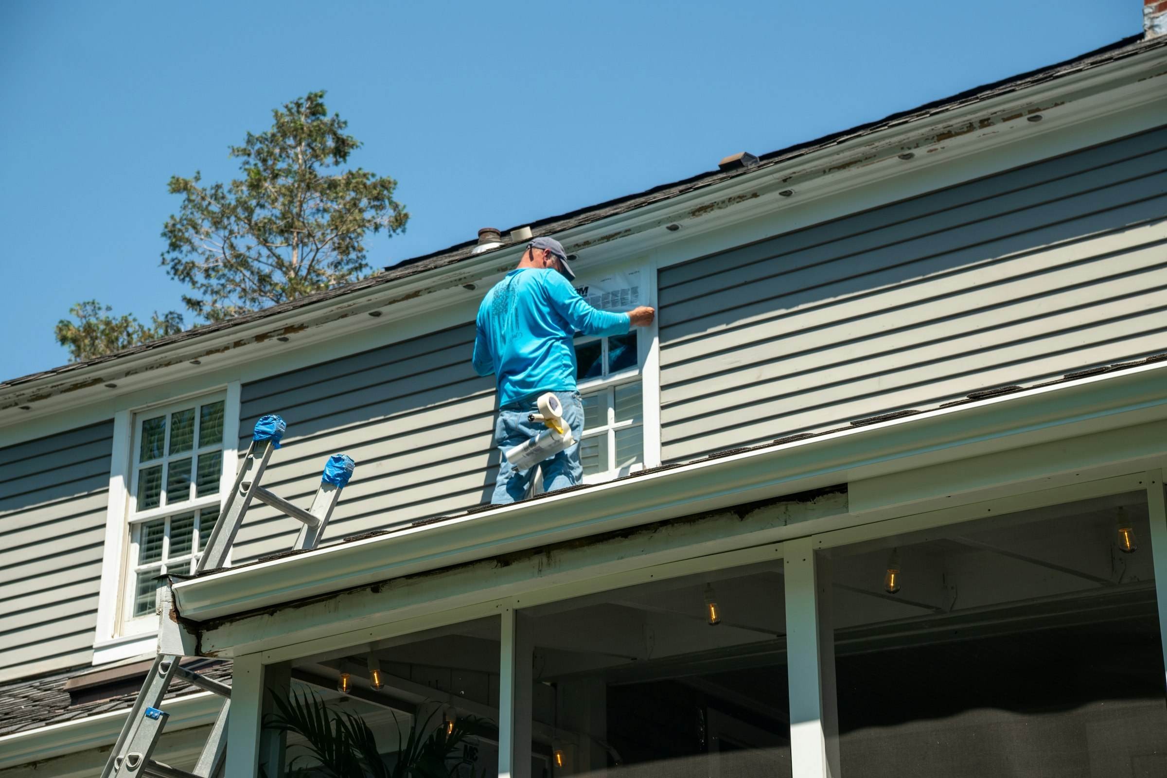 A roofer installing flashing along a window on a grey-sided home